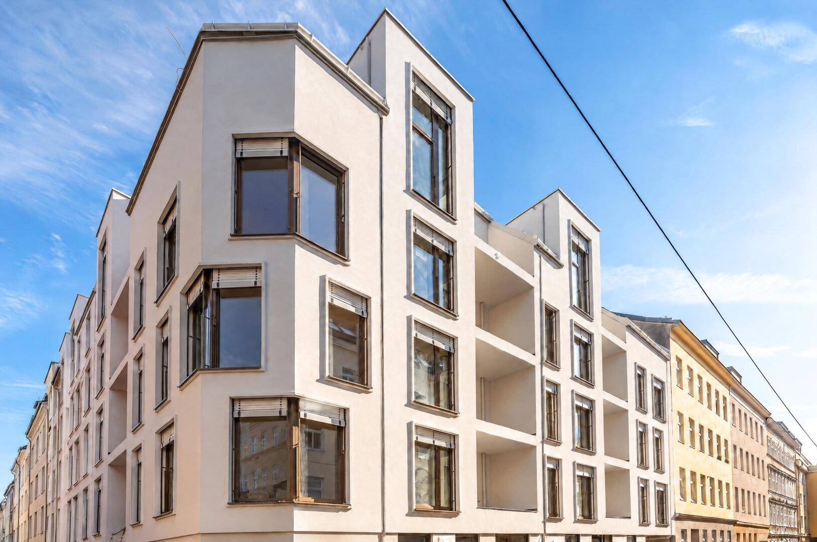Moderne weiße Fassade eines Neubaus mit vielen Fenstern und Balkonen unter blauem Himmel.
