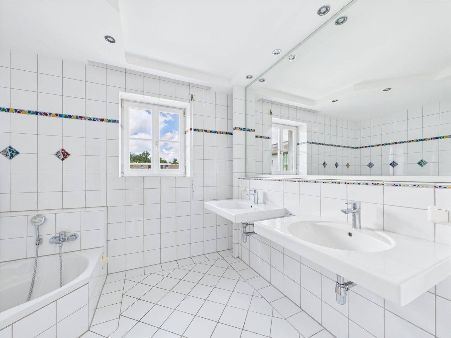Helles Badezimmer mit weißen Fliesen, Doppelwaschbecken und Fenster.