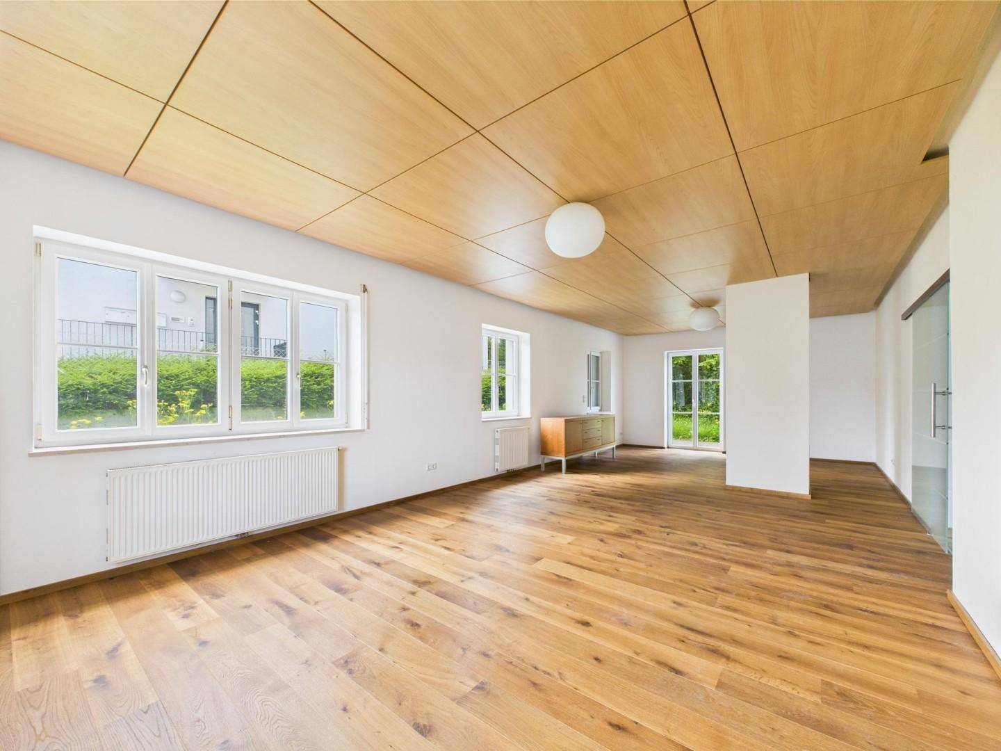 Geräumiges Wohnzimmer mit Holzboden, großen Fenstern und Holzdeckenpaneelen.