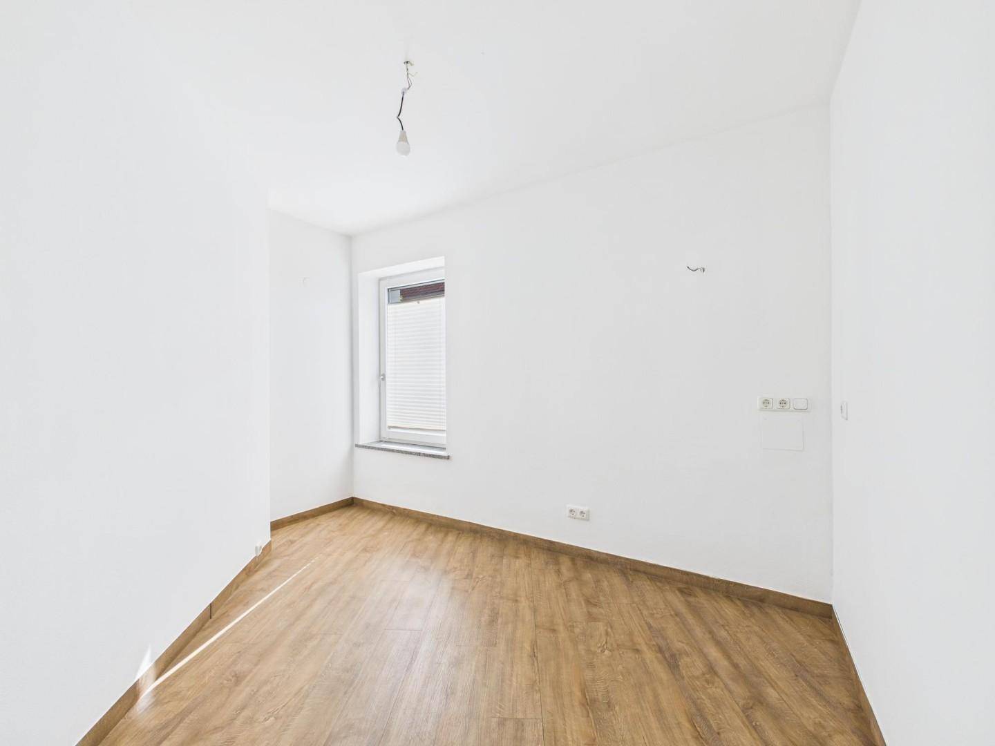 Leeres Zimmer mit Laminatboden und einem Fenster, das viel Tageslicht hereinlässt, ideal für ein Schlafzimmer.