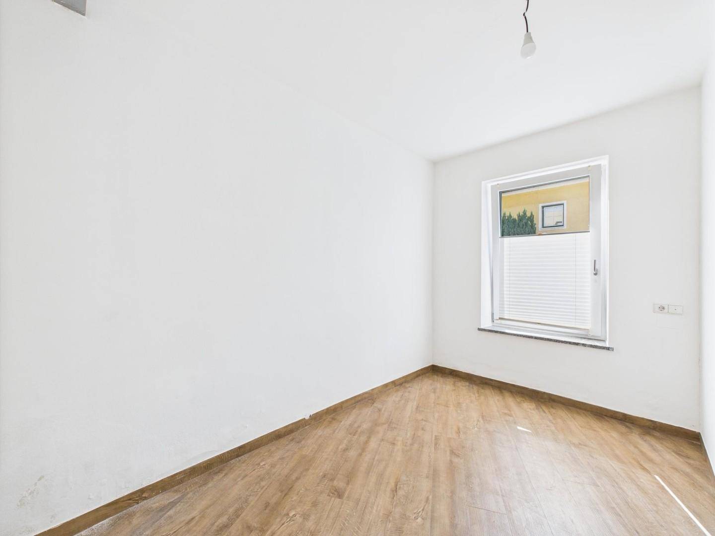 Helles, leeres Zimmer mit Laminatboden und einem Fenster mit Blick ins Grüne, bereit zur Einrichtung.