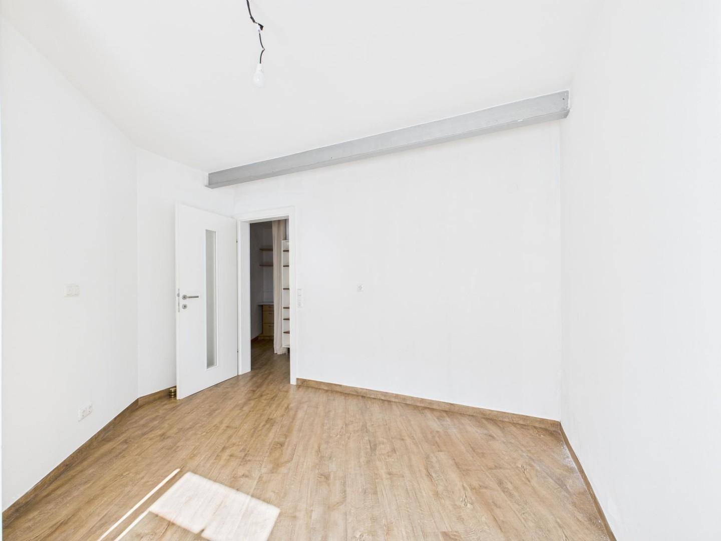Leeres Zimmer mit Laminatboden, weiß gestrichenen Wänden und einer Tür zu einem angrenzenden Nebenraum.