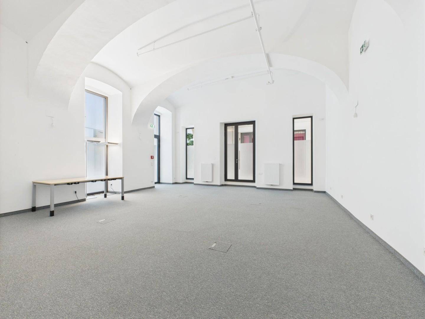 Weitläufiger Büroraum mit hohen Gewölbedecken, großen Fenstern und grauem Teppichboden.