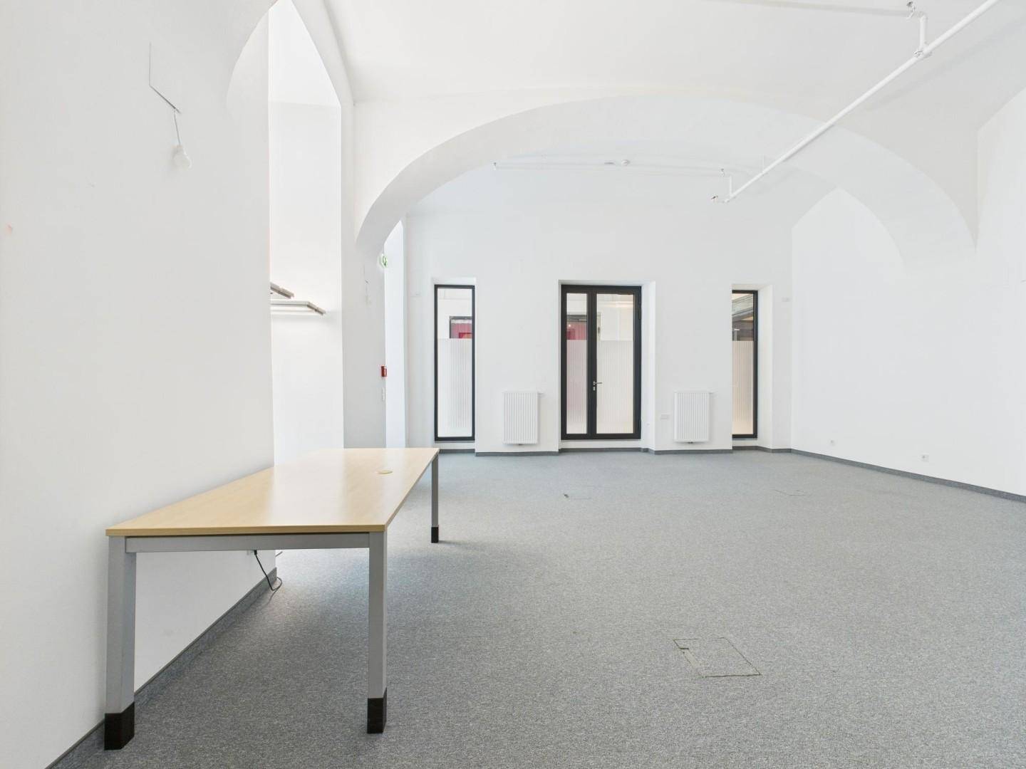 Geräumiger, leerer Büroraum mit Gewölbedecken, großen Fenstern und einem einfachen Tisch.