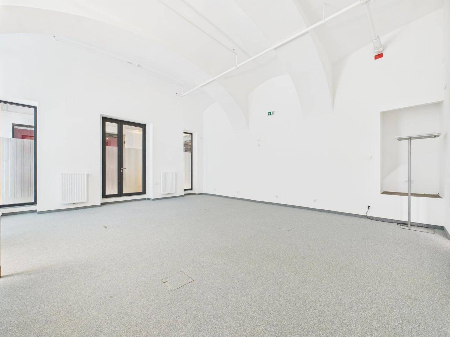 Heller Büroraum mit hohen Gewölbedecken, großen Fenstern und sauberem Teppichboden in Grau.