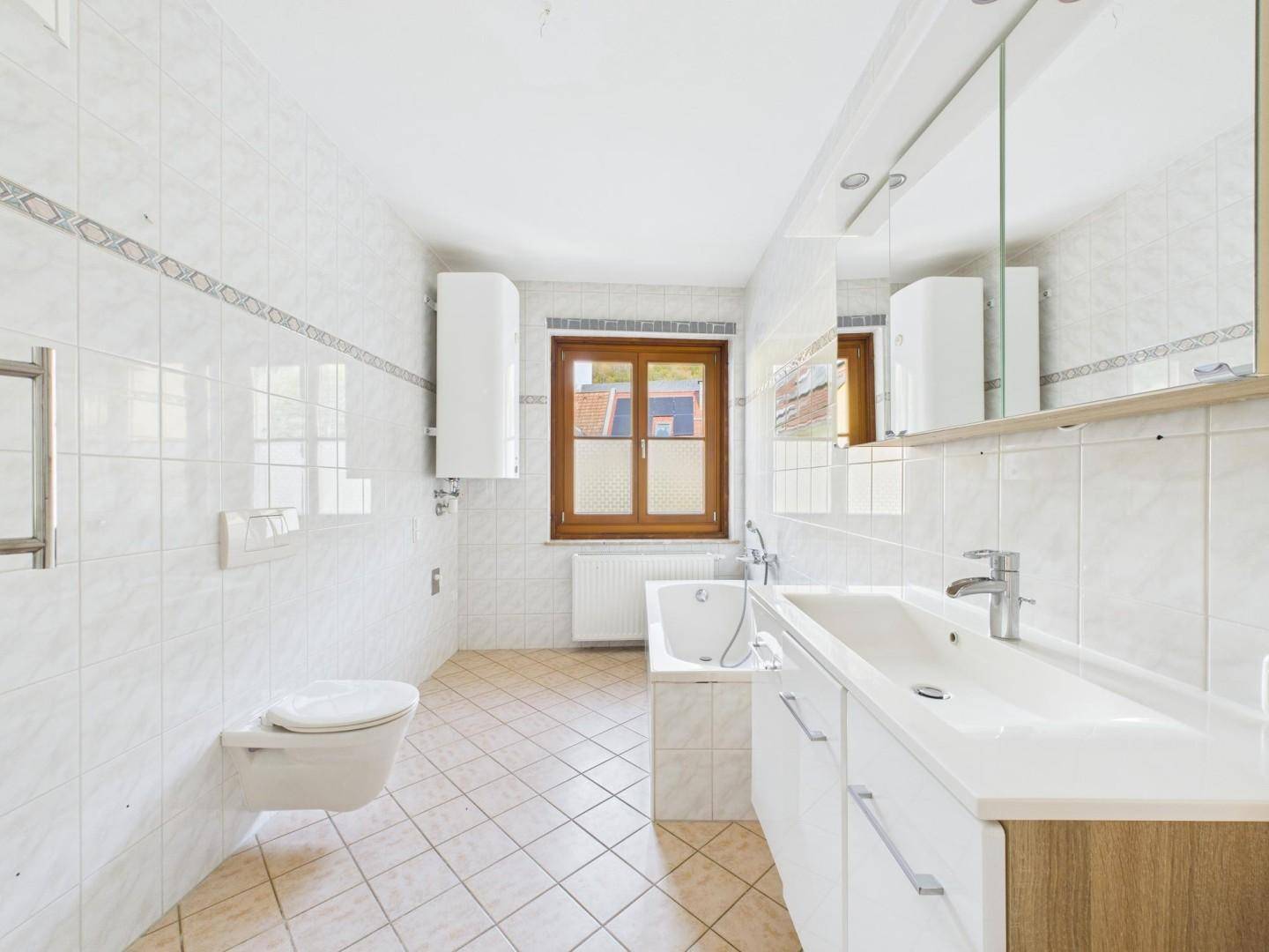 Helles Badezimmer mit weißen Fliesen, Badewanne, WC und großem Waschbeckenunterschrank mit Spiegelschrank.