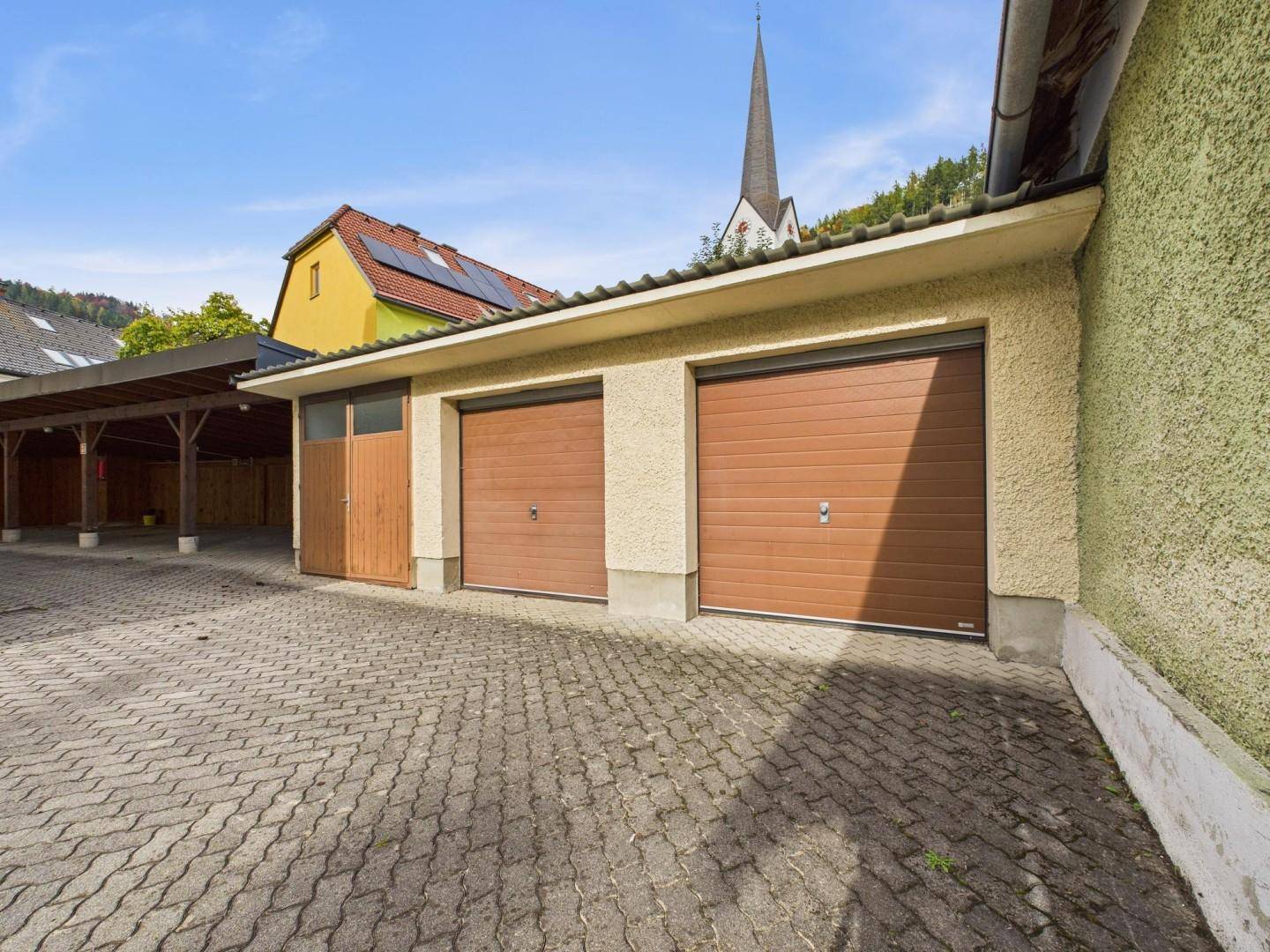Doppelgarage mit braunen Toren und gepflastertem Vorplatz, neben einem überdachten Carportbereich.