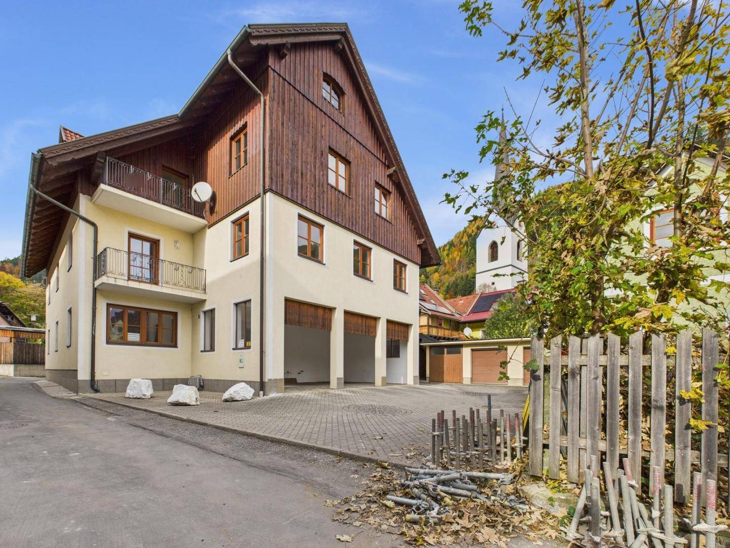 Rückansicht des Gebäudes mit Holzverkleidung, Balkonen und Garagenzugang, umgeben von Bäumen und einem Kirchturm.