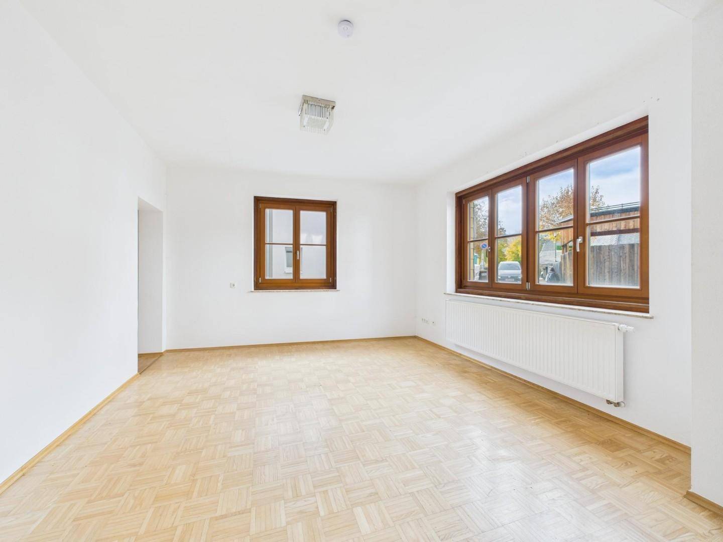 Lichtdurchfluteter Raum mit großen Holzfenstern, Parkettboden und Heizkörper, ideal für Wohnzwecke.