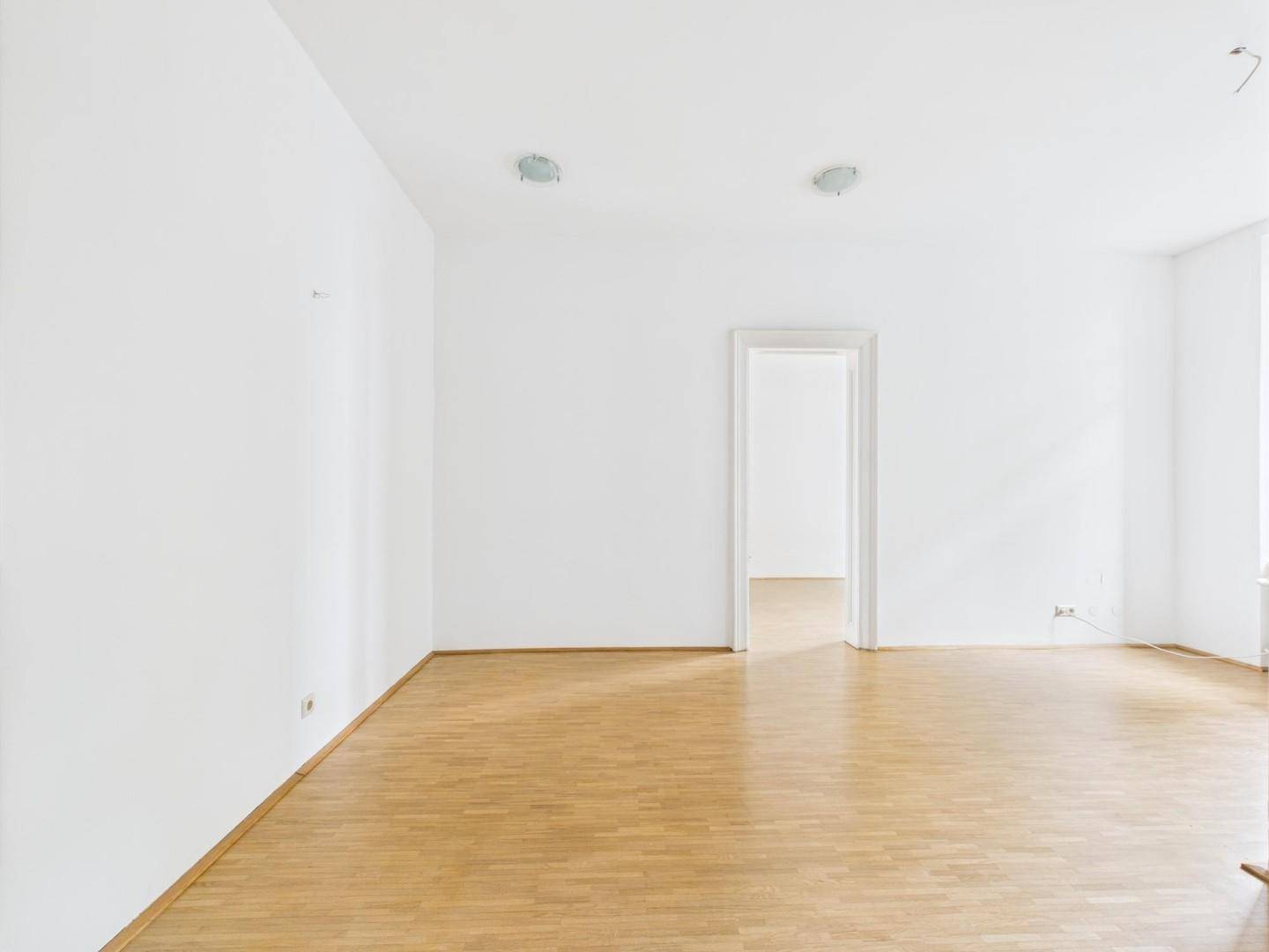 Heller, offener Raum mit Parkettboden und weißen Wänden, verbunden durch eine breite Türöffnung.