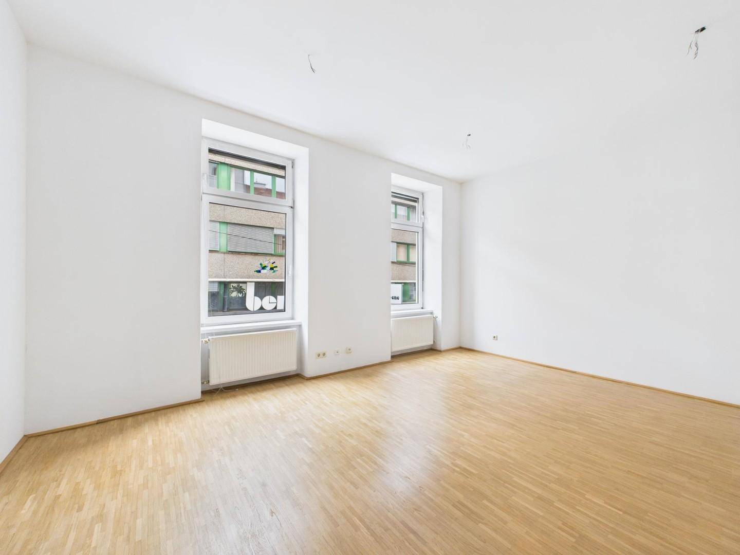 Heller Wohnraum mit Parkettboden, weißen Wänden und zwei Fenstern mit Blick auf die Stadt.