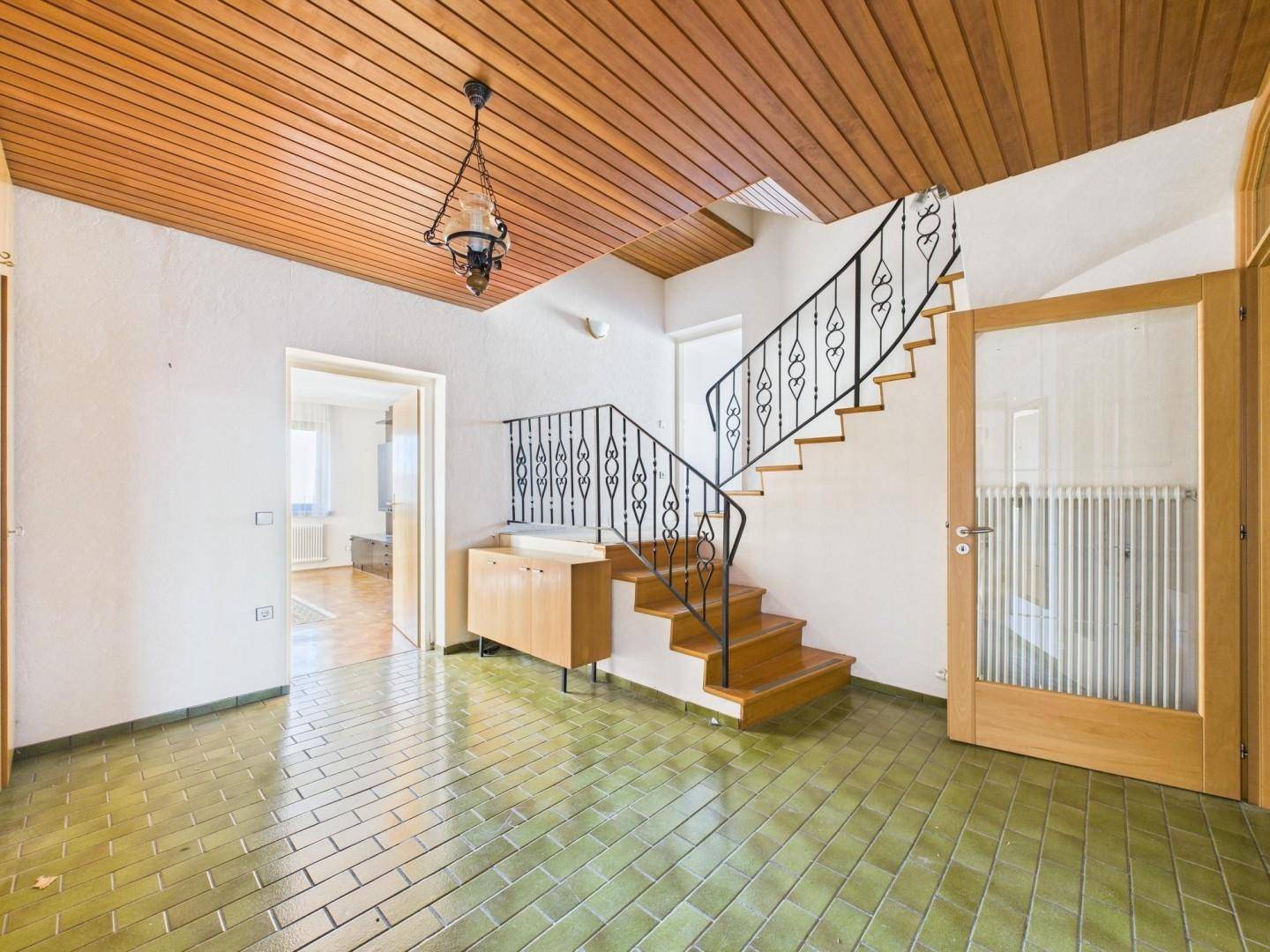 Heller Flur mit Holztreppe, schmiedeeisernem Geländer und grünen Bodenfliesen.