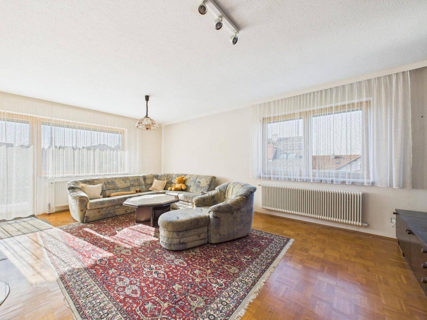 Geräumiges Wohnzimmer mit Parkettboden, großem Teppich und viel Tageslicht durch die Fenster.