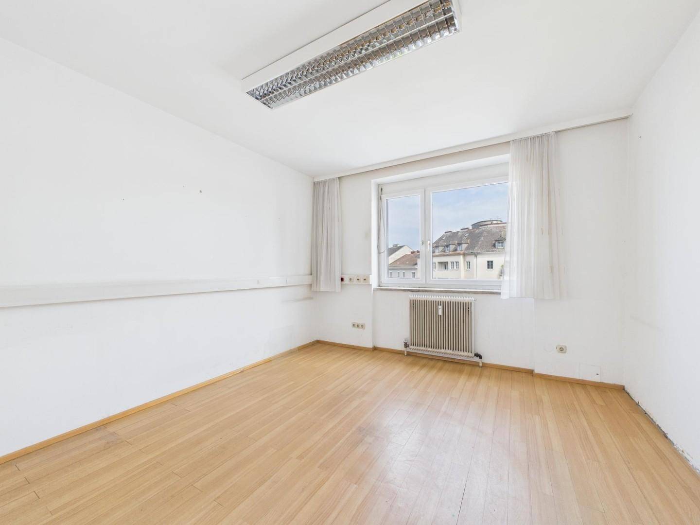 Helles, leeres Büro mit Laminatboden, großem Fenster und Blick auf die umliegenden Gebäude.