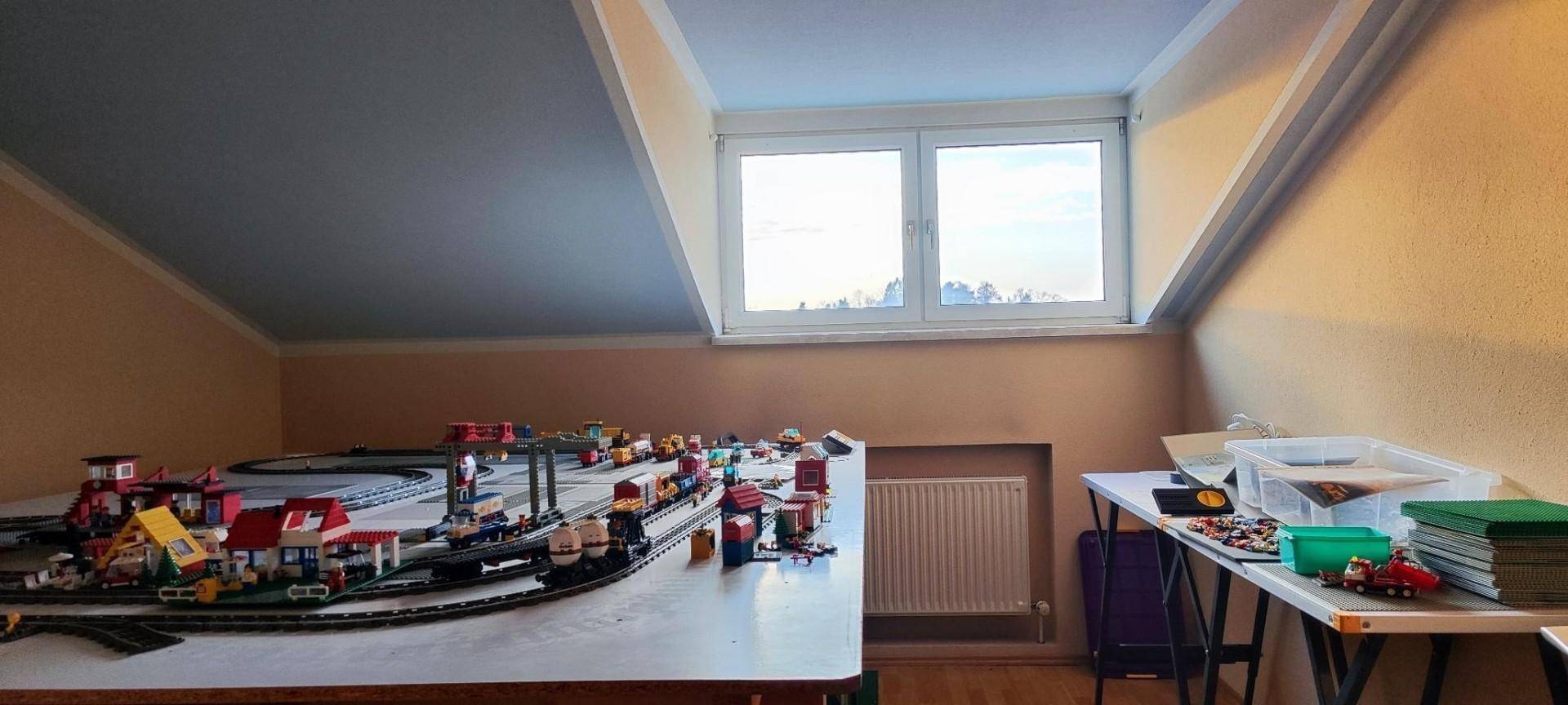 Dachzimmer mit schräger Decke, großem Fenster und einem Spielzeugzug auf dem Tisch.