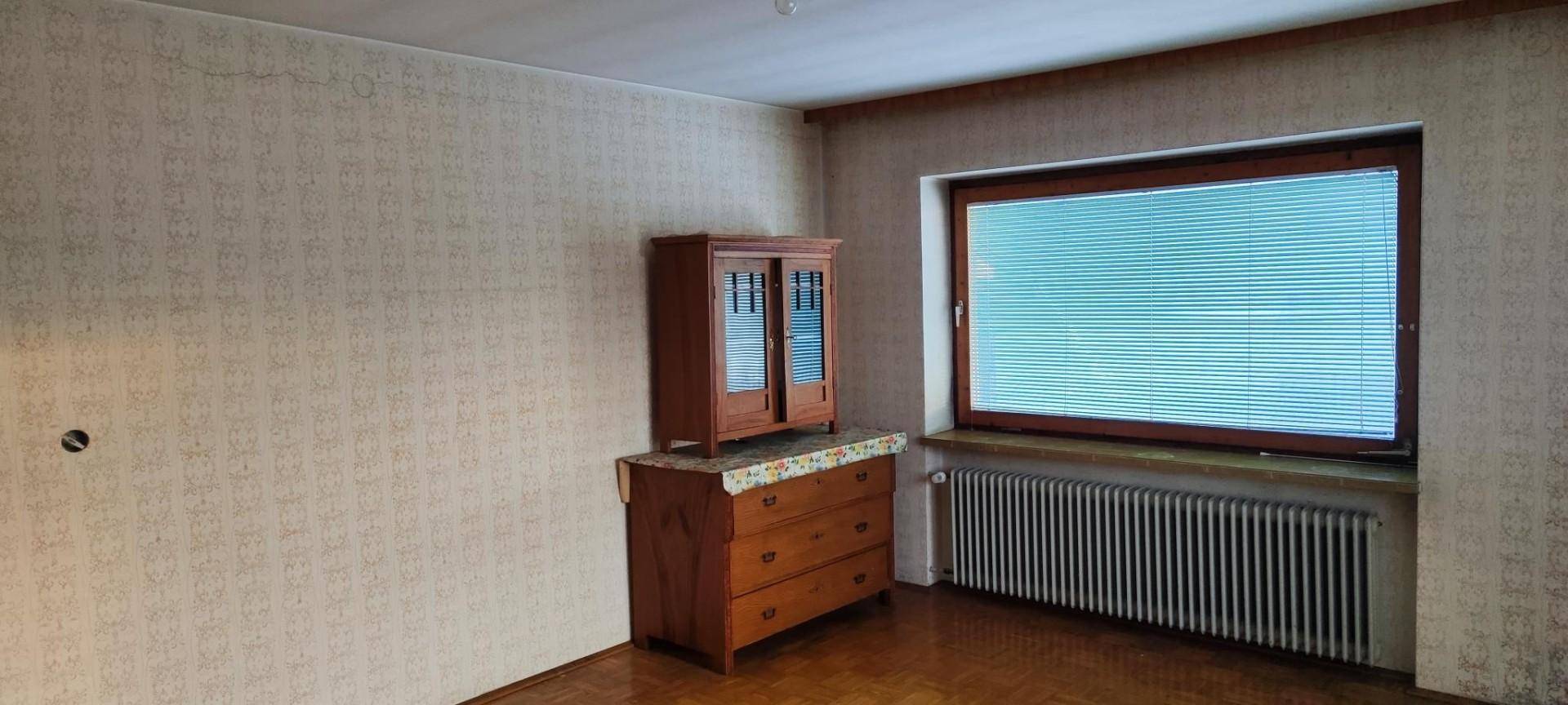 Zimmer mit gemusterter Tapete, Holzschrank und großem Fenster mit Jalousien.