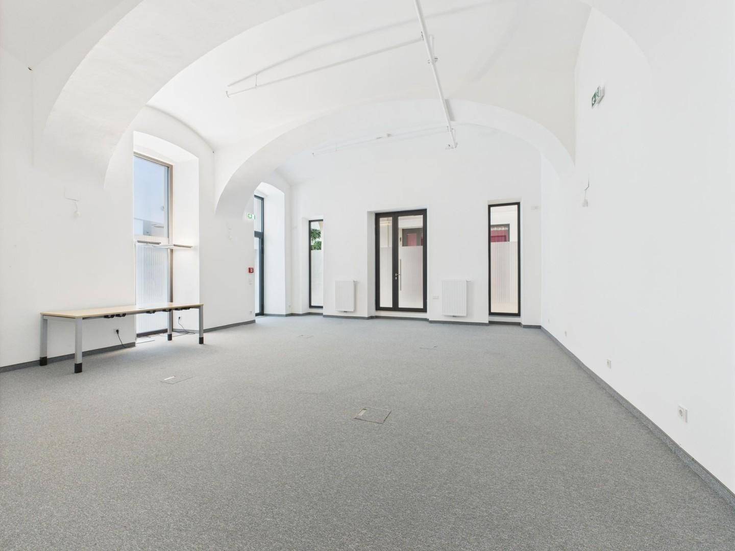 Heller Gewerberaum mit hohen, gewölbten Decken, großen Fenstern und grauem Teppichboden.