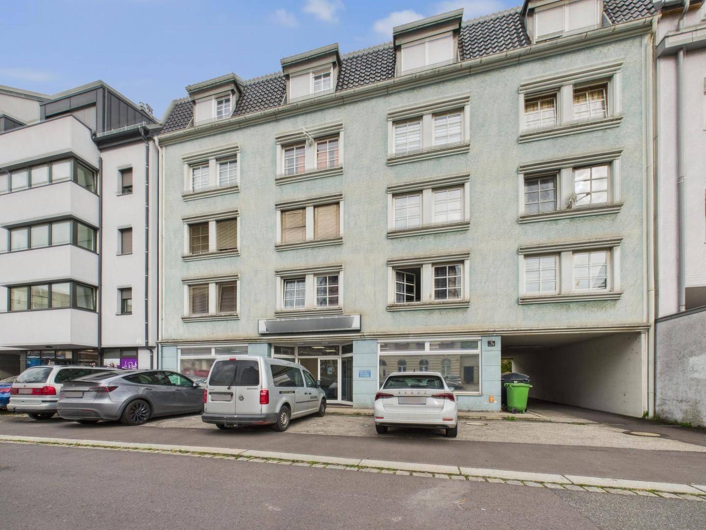 Mehrstöckiges Gebäude mit Geschäftsräumen im Erdgeschoss und Wohnungen darüber, inklusive Parkplätzen und überdachtem Bereich.