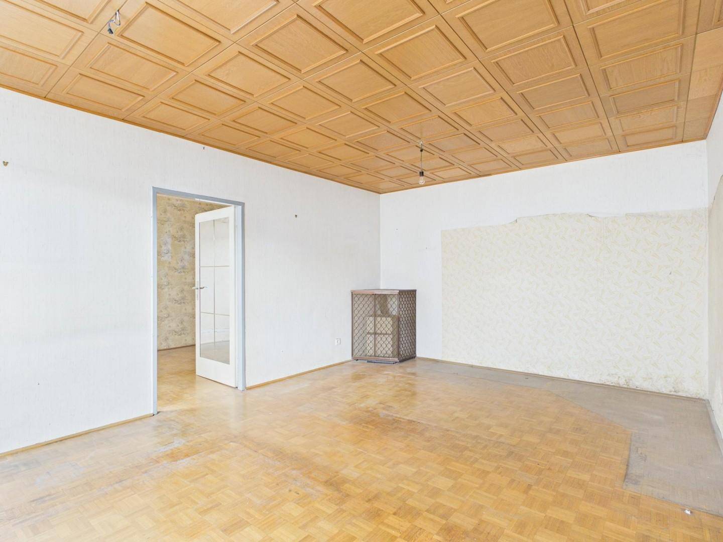 Leeres Wohnzimmer mit Fischgrätparkettboden und einer Holzpaneeldecke, bereit zur Renovierung.