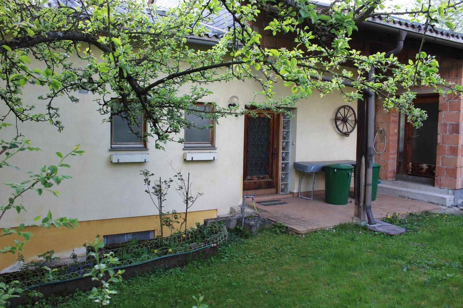 Seitenansicht des Hauses mit Eingangsbereich, kleiner gepflasterter Fläche und blühenden Bäumen.
