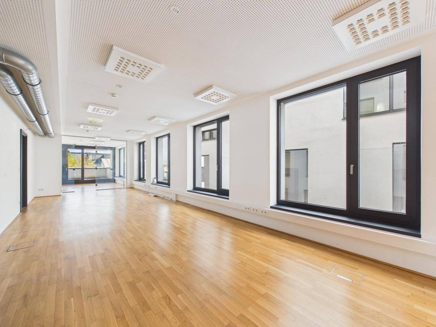 Geräumiger Bürobereich mit hellem Holzboden, großen Fenstern und moderner Deckenbeleuchtung.