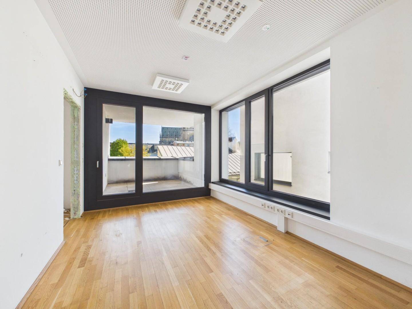 Heller Büroraum mit Holzboden, großen Fenstern und Zugang zu einem kleinen Balkon.