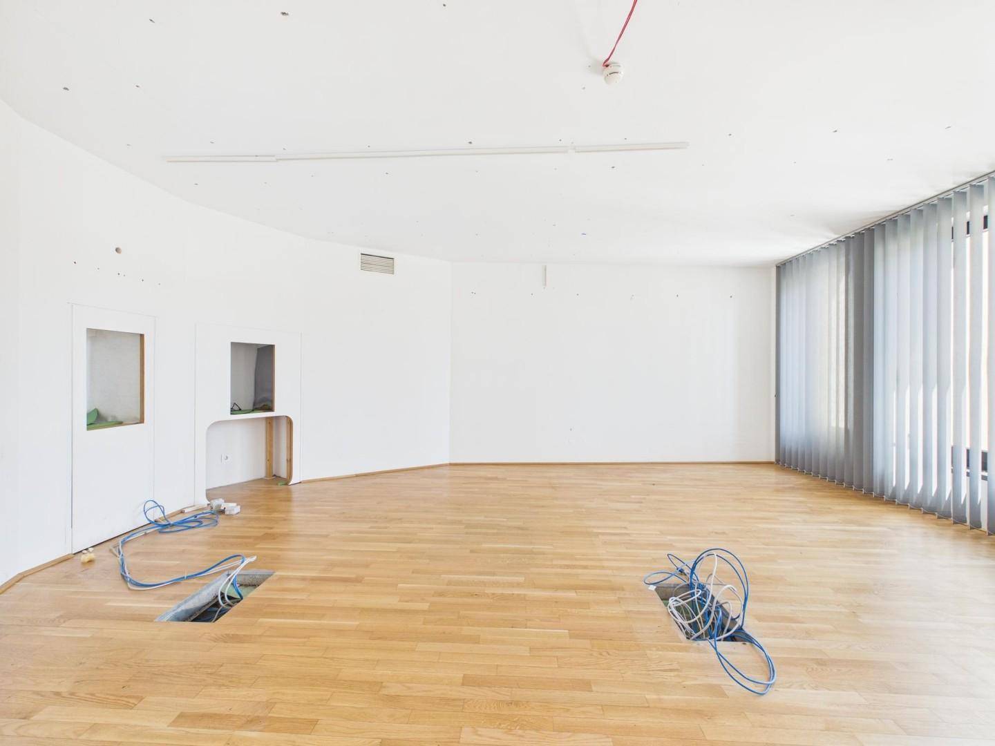 Großer, leerer Büroraum mit Holzboden, weißen Wänden und sichtbaren Kabeln für die Installation.