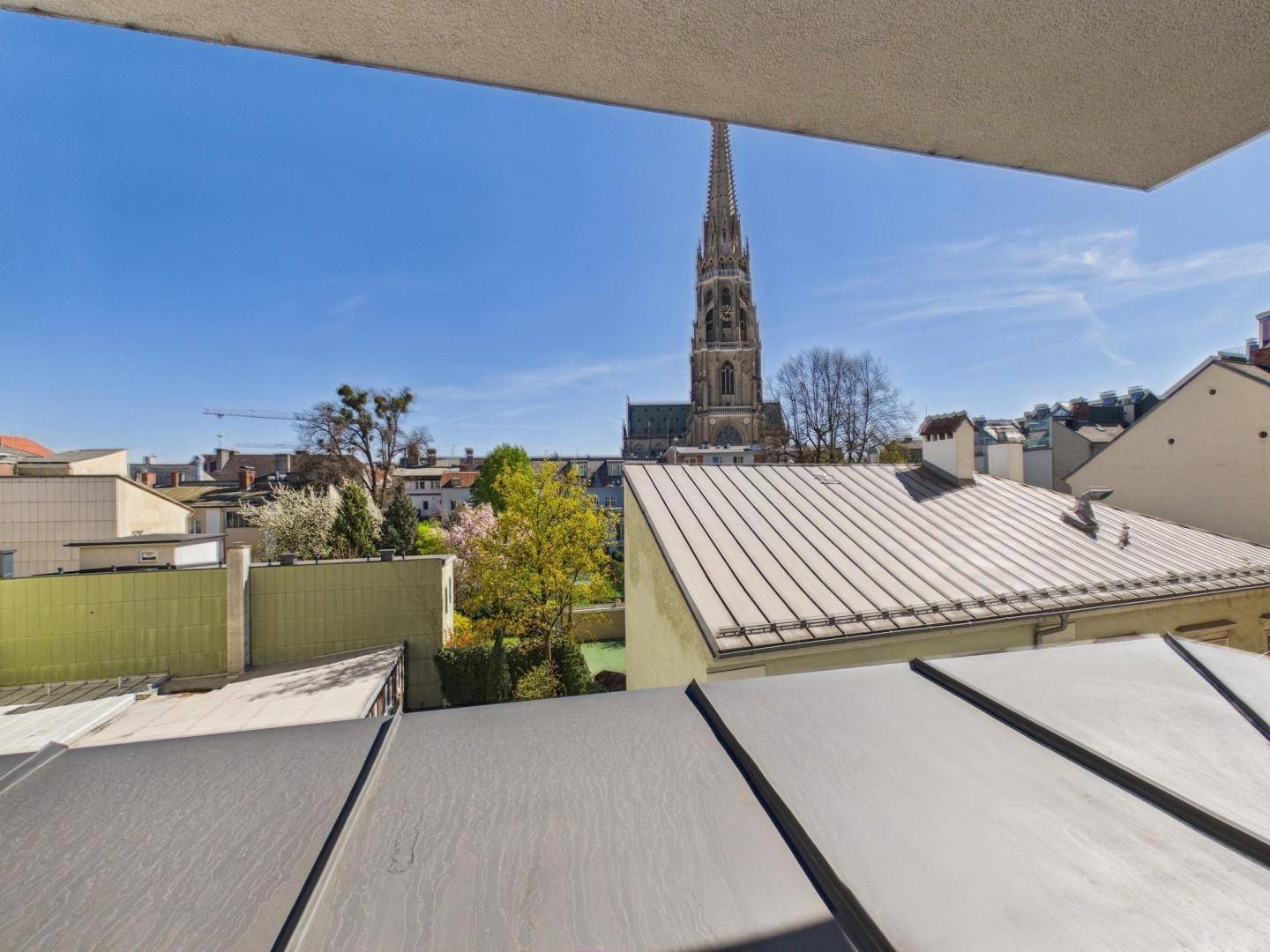 Balkon mit weitem Blick über die Stadt, Dächer und einen markanten Kirchturm unter blauem Himmel.