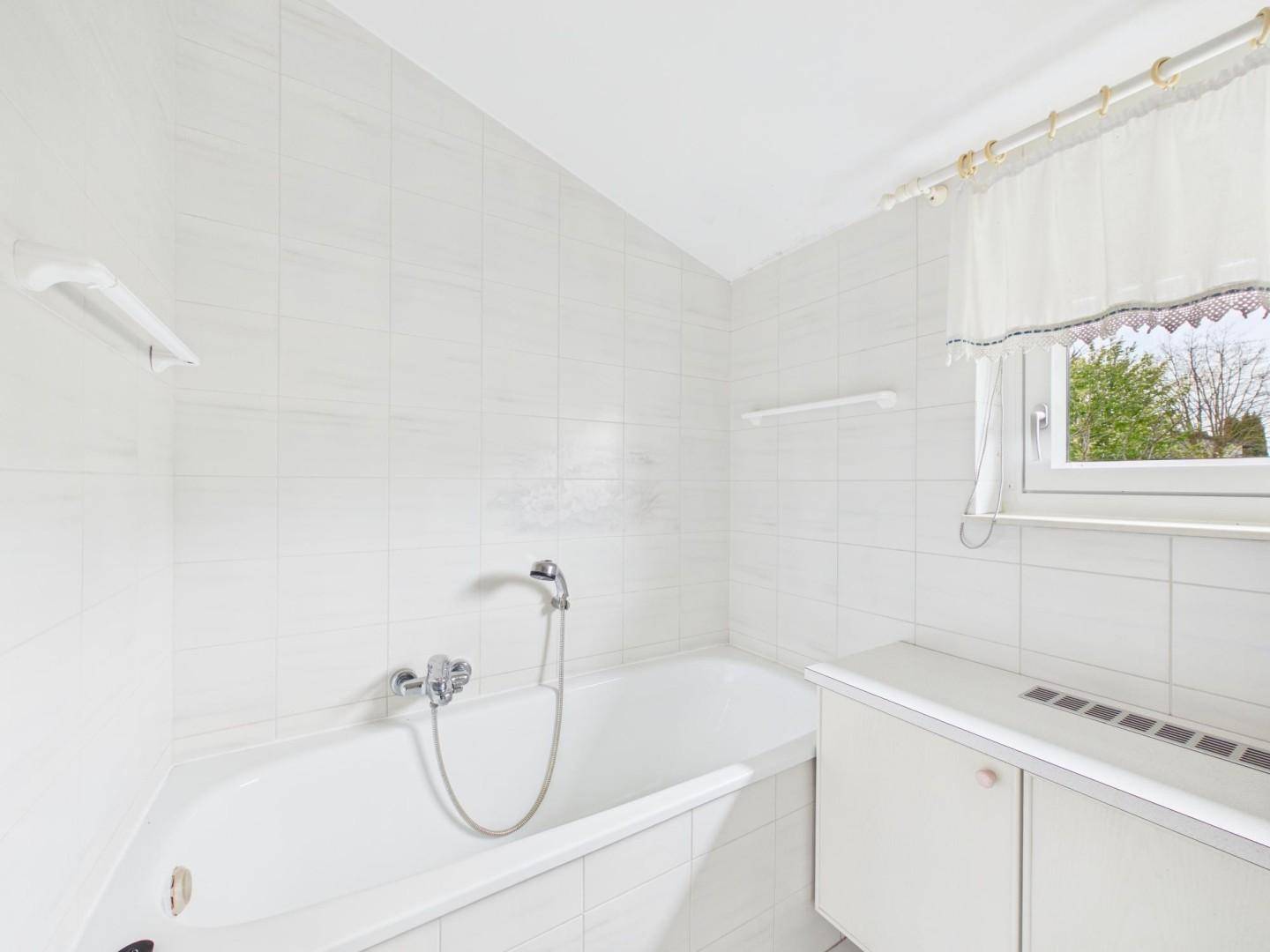 Helles Badezimmer mit weißen Fliesen, Badewanne und einem Fenster für Tageslicht.