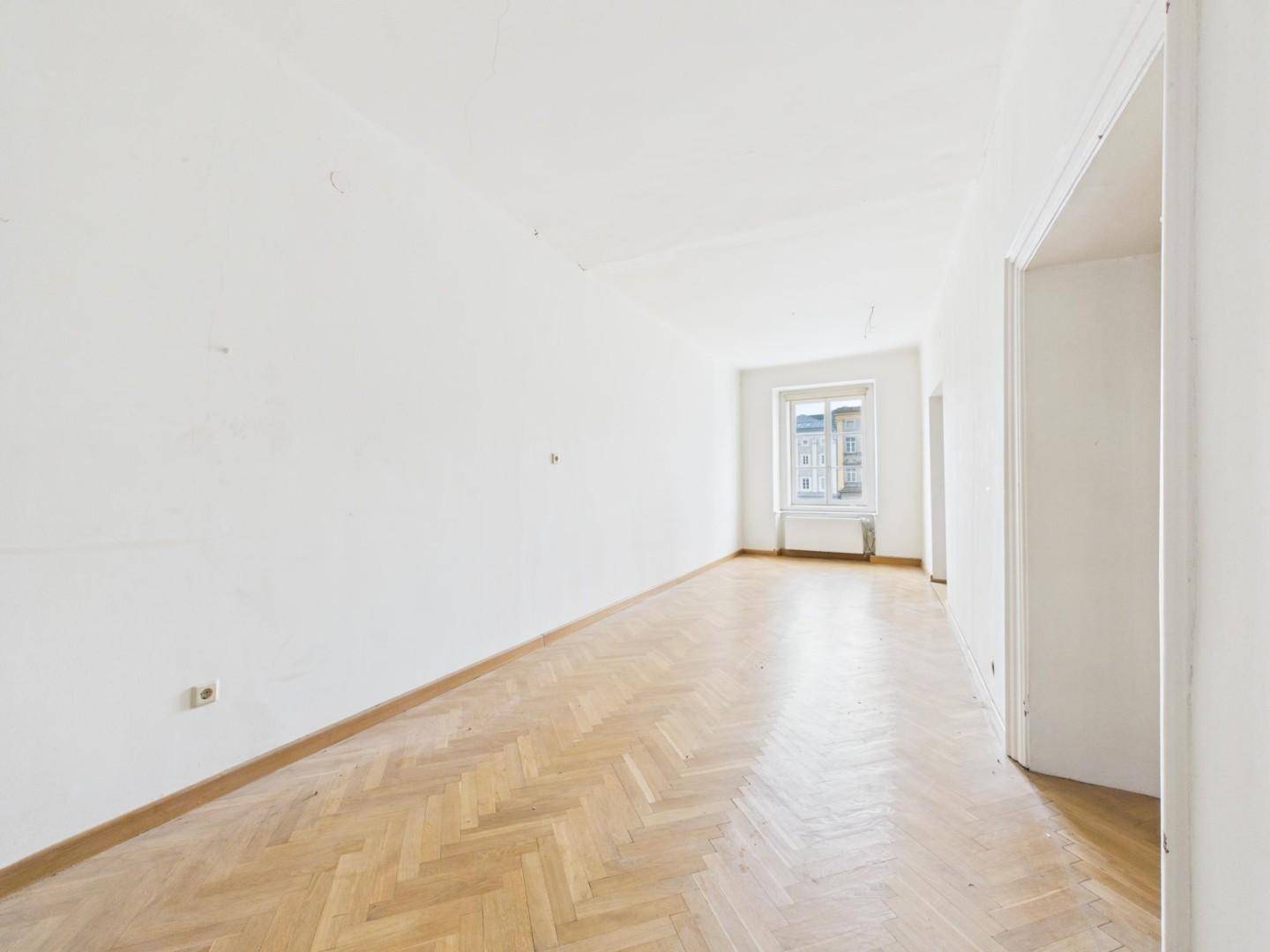 Langer, heller Raum mit Fischgrätparkett und einem Fenster am Ende, das für natürliche Belichtung sorgt.