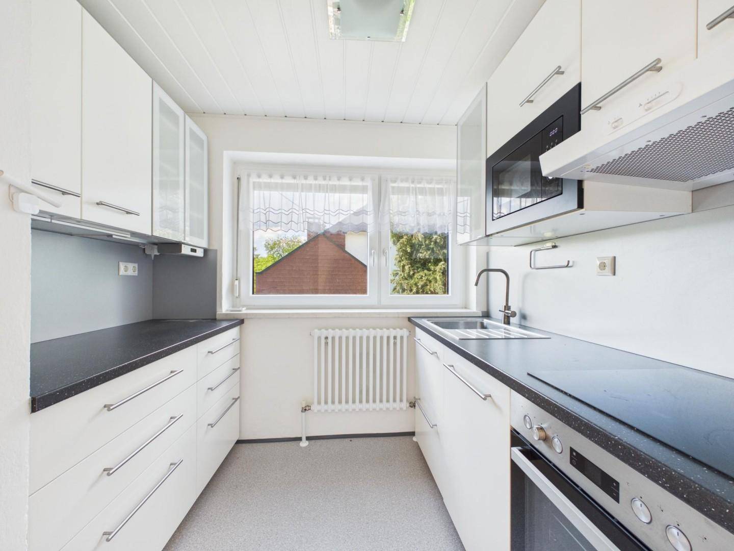 Funktionale Küche mit weißen Schränken, dunkler Arbeitsplatte und Fenster mit Blick nach draußen.
