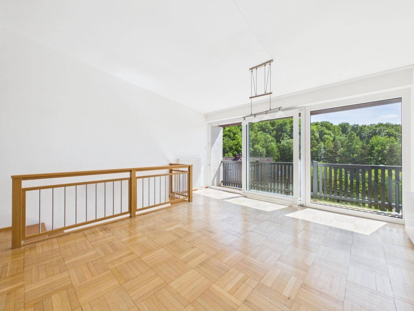 Heller Wohnbereich mit Fischgrätparkett und breitem Zugang zu einem Balkon mit grüner Aussicht.