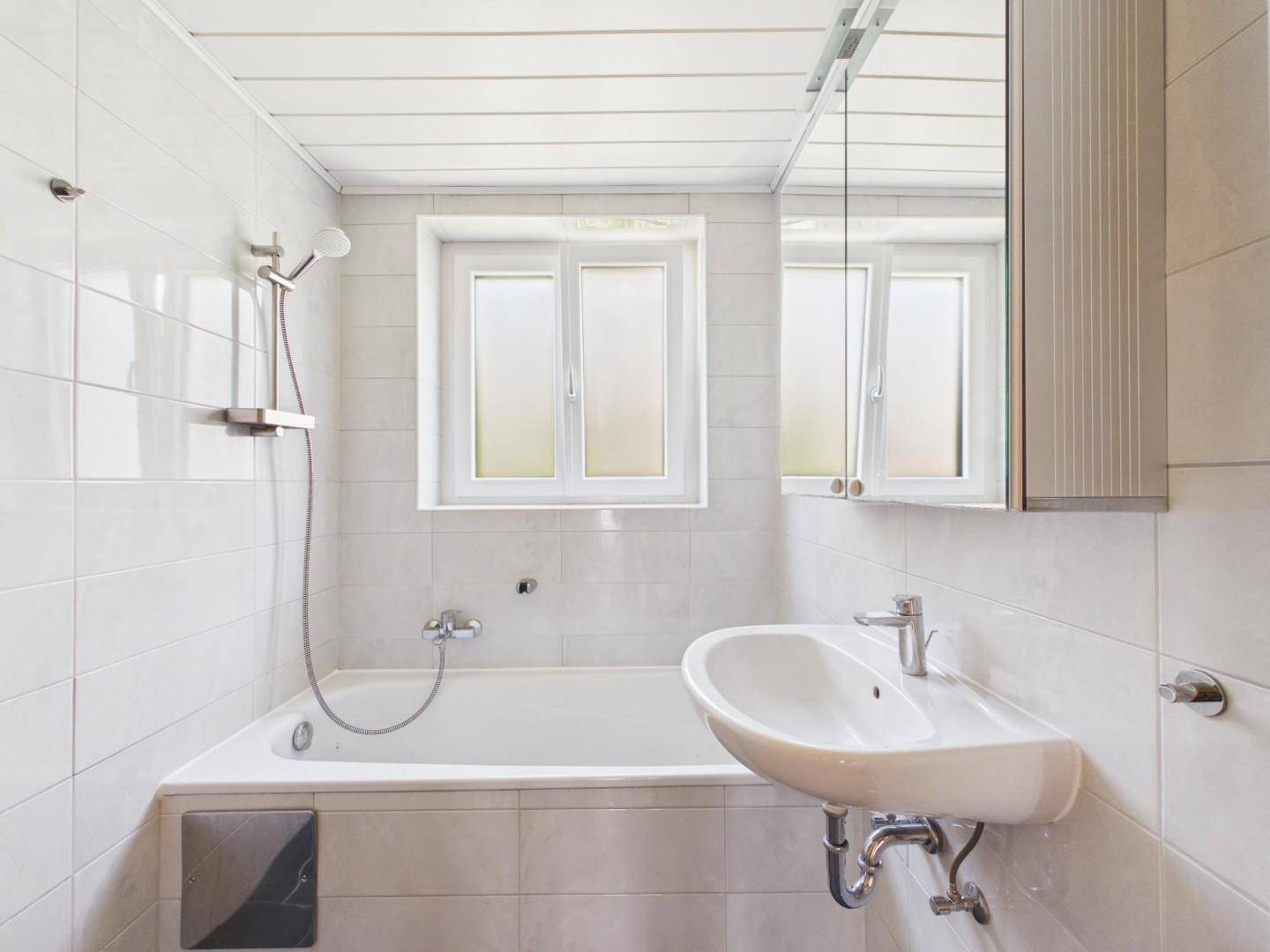 Helles Badezimmer mit weißen Fliesen, Badewanne mit Duschfunktion, Waschbecken und Fenster.