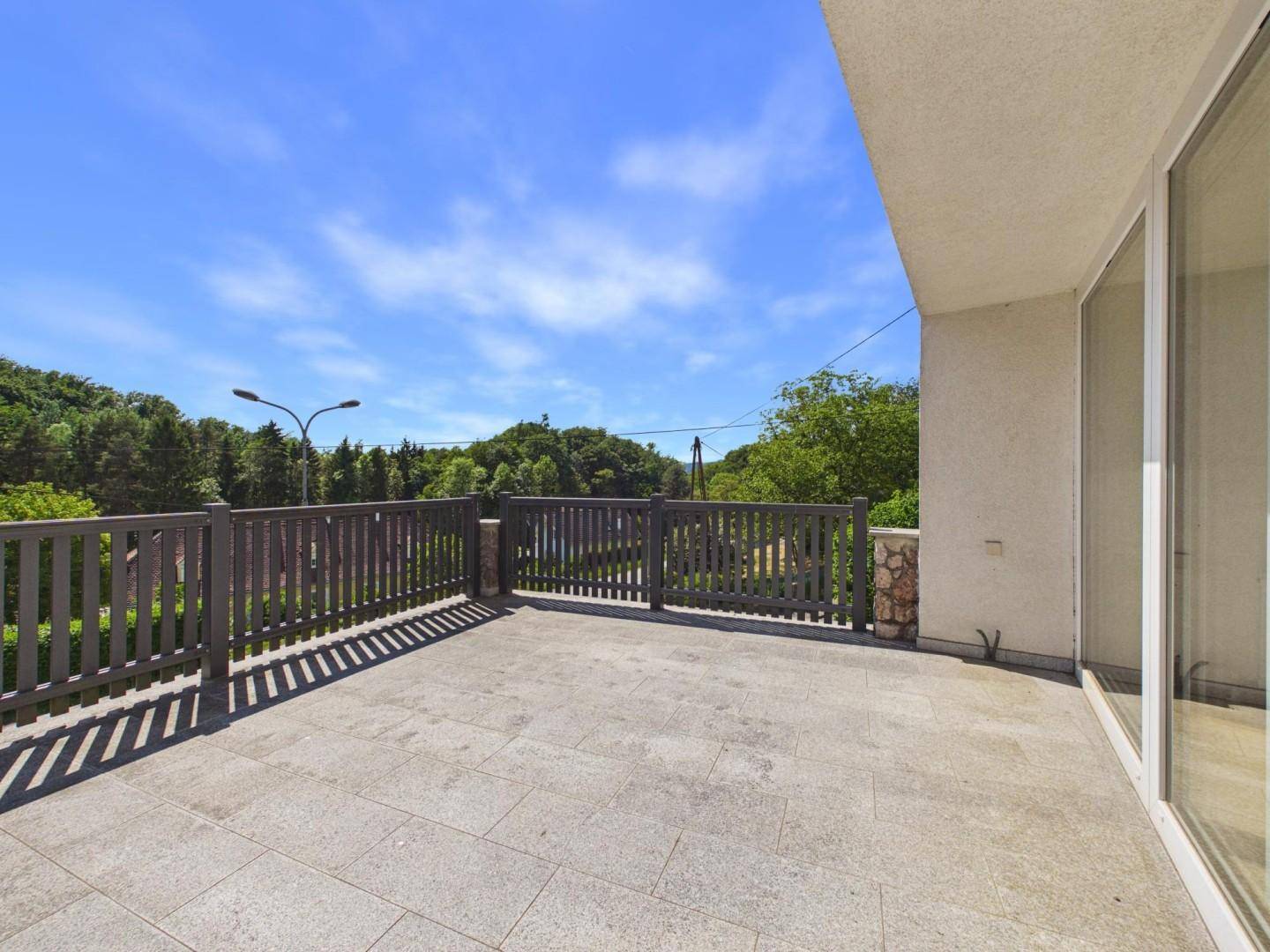Große gepflasterte Terrasse mit Holzgeländer und Blick ins Grüne unter blauem Himmel.