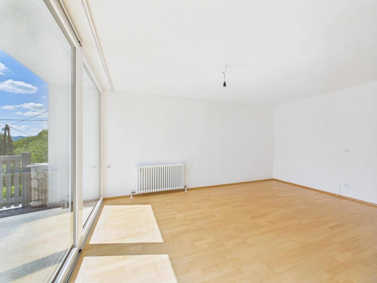 Heller, leerer Raum mit Parkettboden, Heizkörper und Zugang zur Terrasse durch große Schiebetüren.