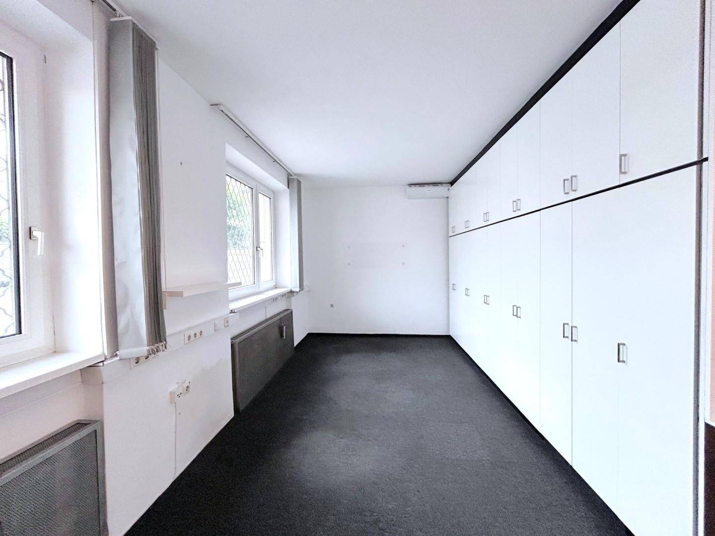 Langer Raum mit dunklem Teppichboden, Einbauschränken und einem Fenster mit Blick ins Grüne.