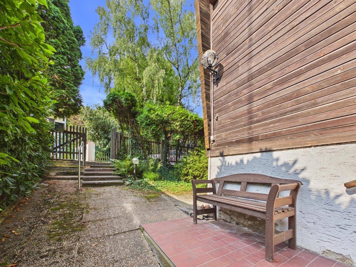 Seitlicher Zugang zum Haus mit Holzbank und gepflastertem Weg, umgeben von Grün.