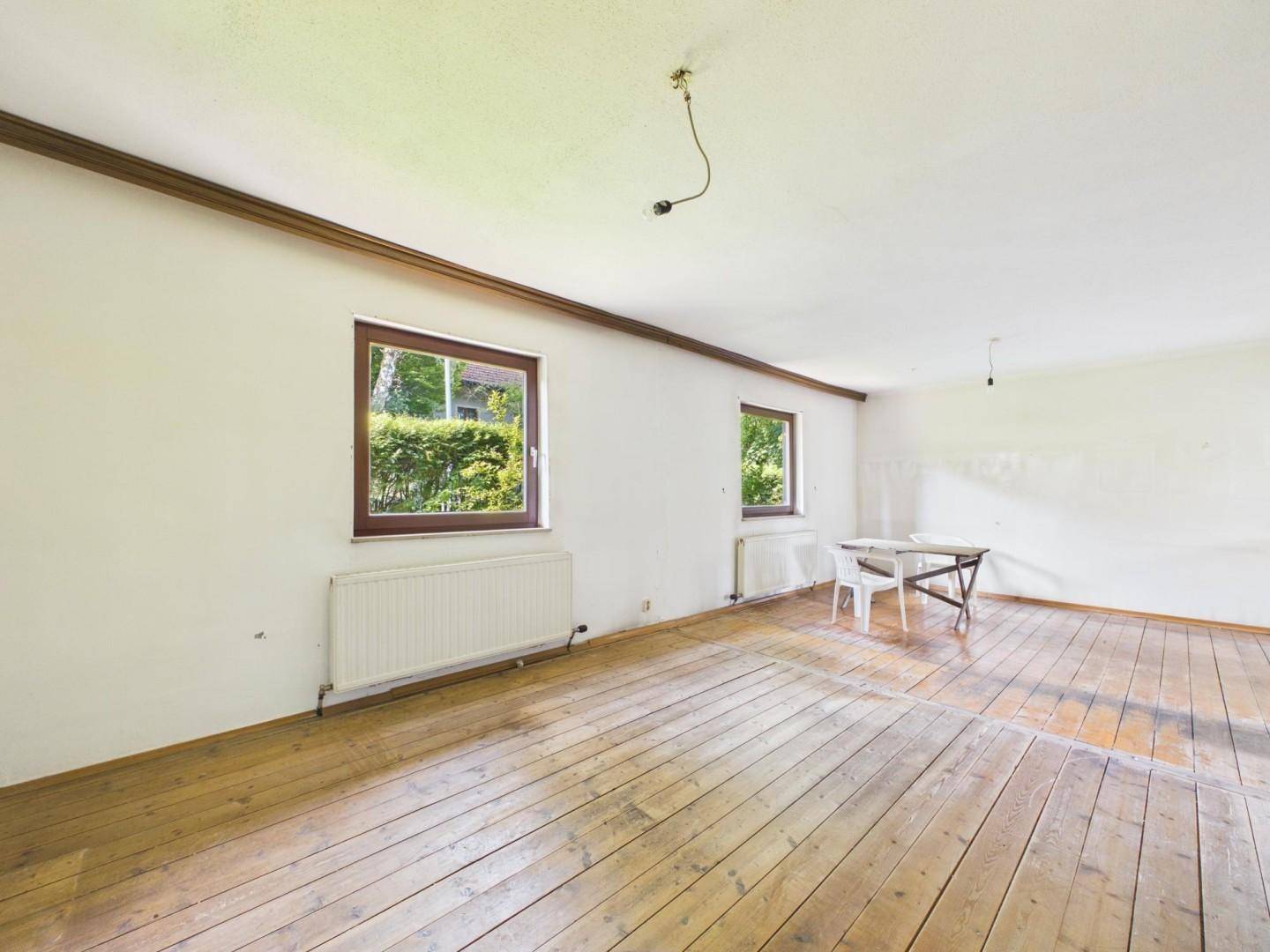 Geräumiges Wohnzimmer mit Holzdielenboden und zwei Fenstern, die viel Tageslicht hereinlassen.