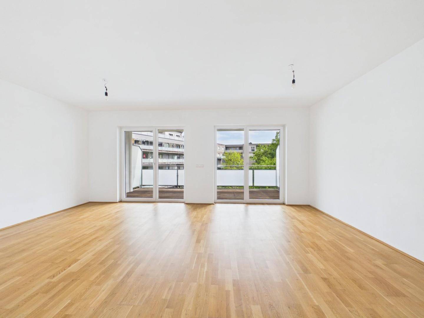 Helles, leeres Wohnzimmer mit Parkettboden und Zugang zu zwei Balkonen.