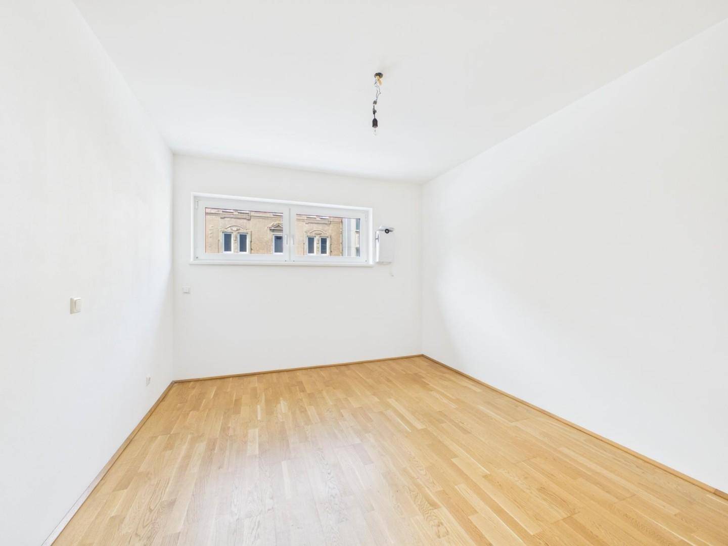Heller Raum mit Parkettboden und Fenster, das auf ein gegenüberliegendes Gebäude blickt.