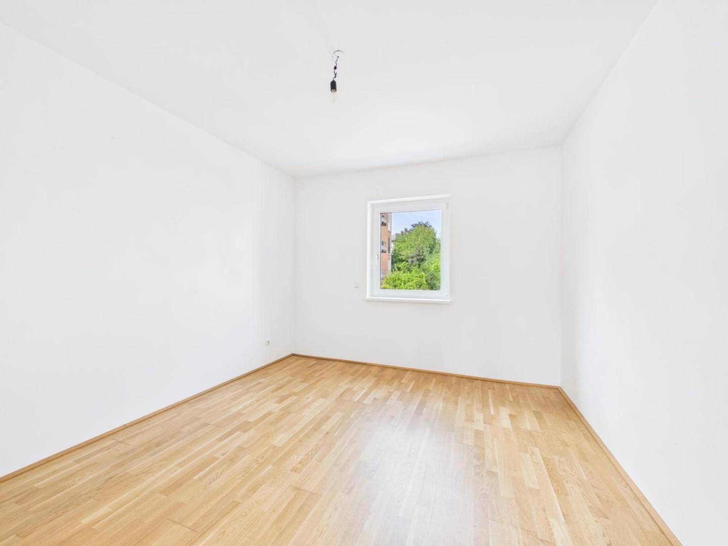 Helles Schlafzimmer mit Parkettboden und Fenster mit Blick ins Grüne.