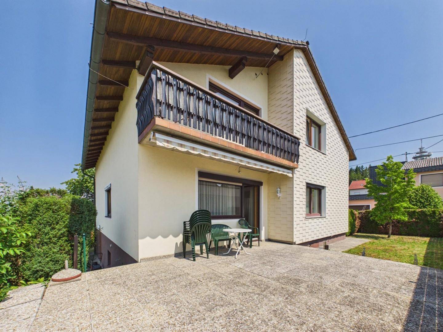 Seitliche Ansicht des Hauses mit Balkon, Terrasse und Gartenmöbeln unter strahlendem Sonnenschein.