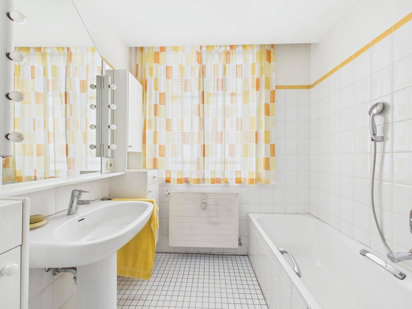 Helles Badezimmer mit Badewanne, Waschbecken und gelb-weiß gemusterten Vorhängen.