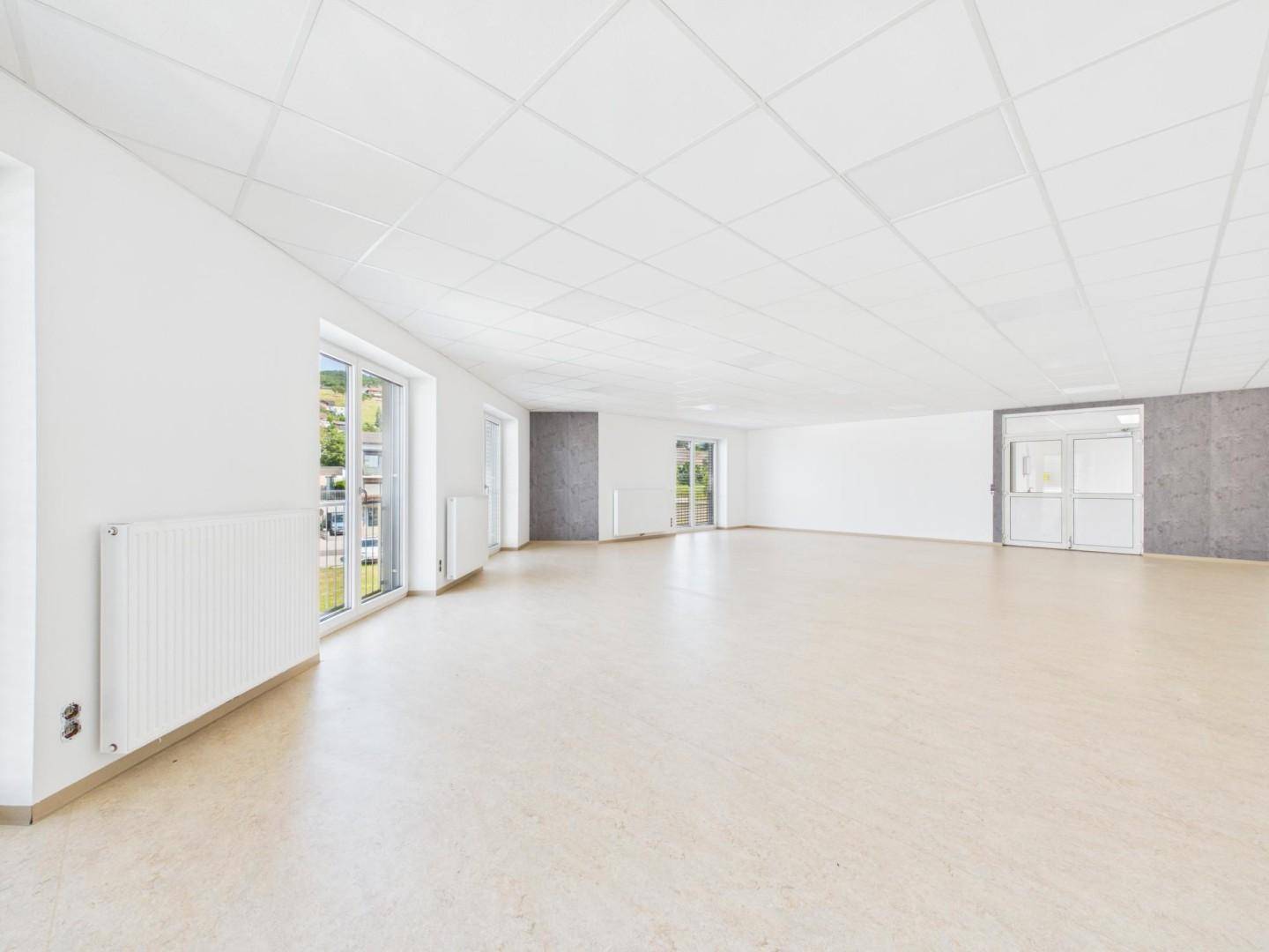 Großer, heller Raum mit hellem Boden, Rasterdecke und Heizkörpern unter den Fenstern, bereit zur Nutzung.