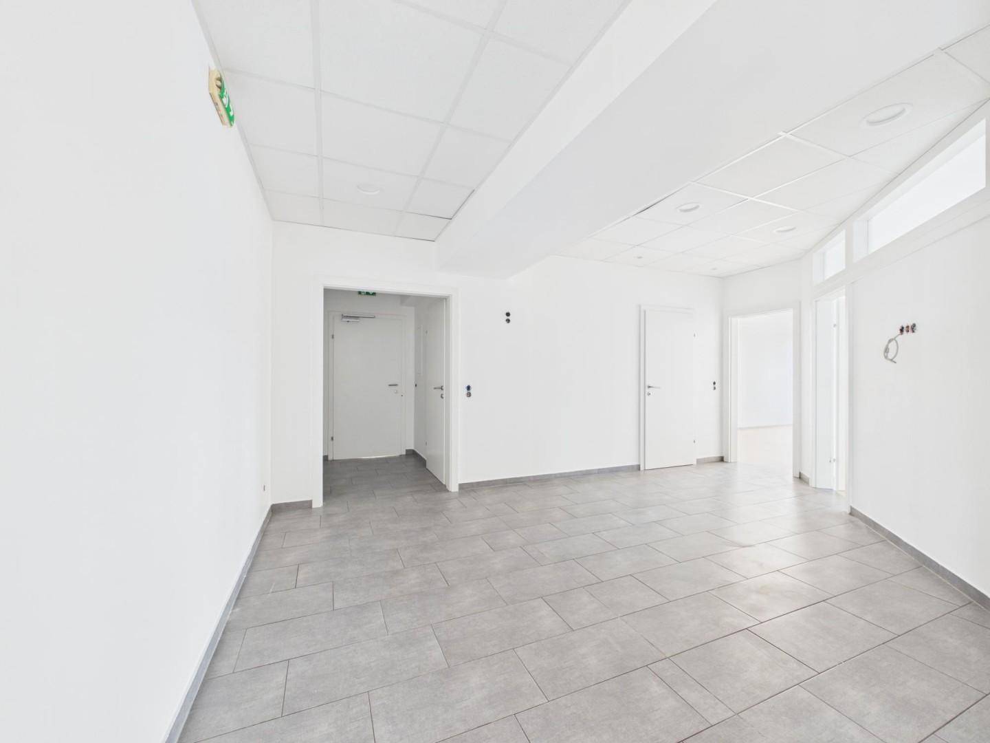 Heller Flur mit grauen Fliesen, weißen Wänden und mehreren Türen, ausgestattet mit Rasterdecke.
