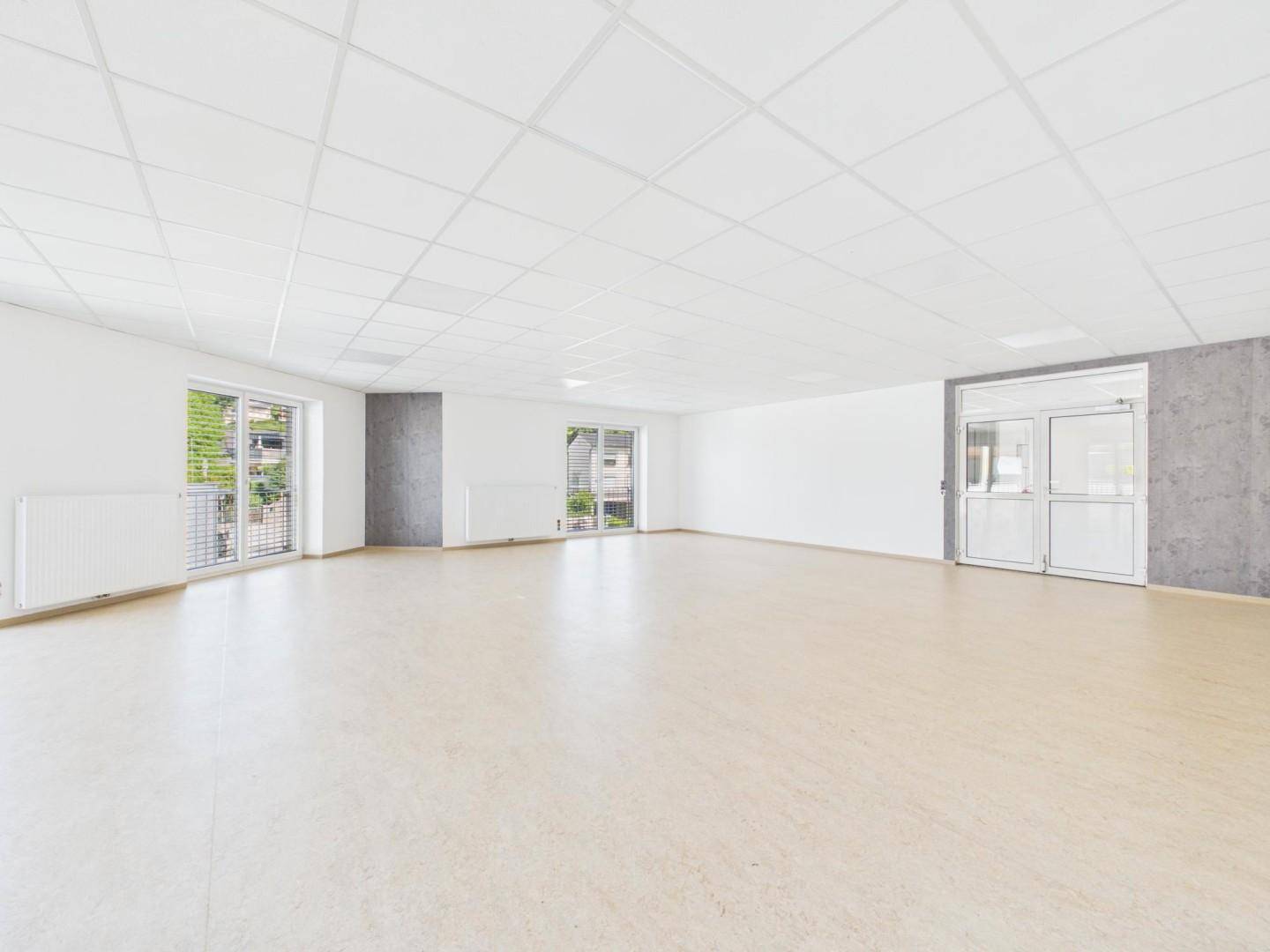Geräumiger, heller Raum mit hellem Boden, Rasterdecke und mehreren Fenstern, inklusive Doppeltür.
