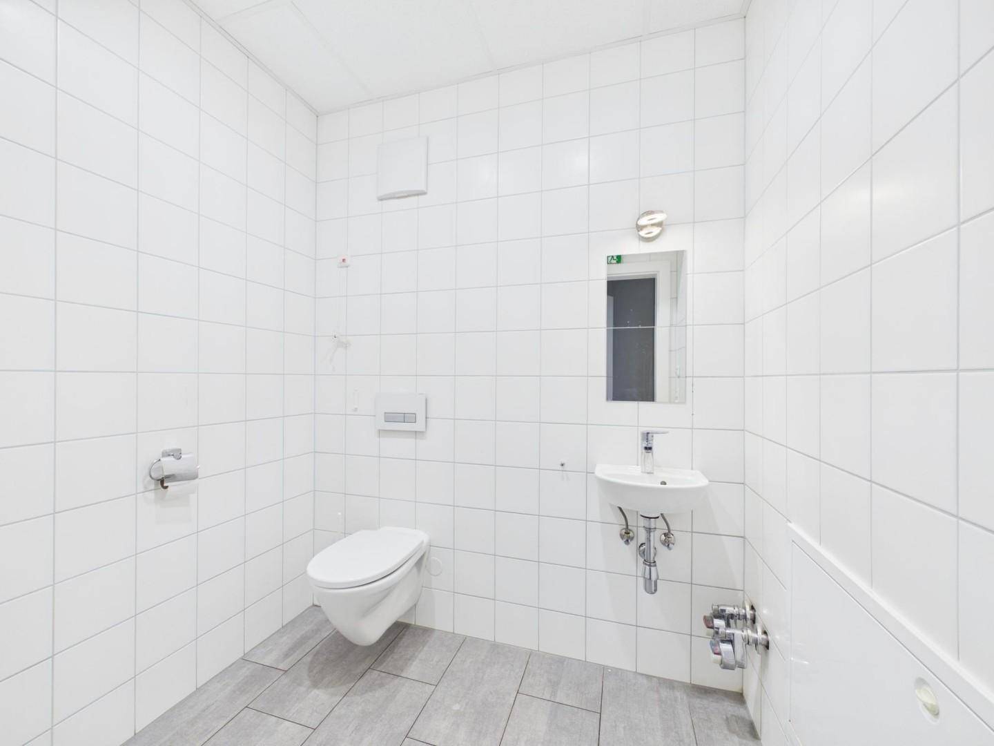 Kleiner, gefliester Toilettenraum mit Wand-WC, Waschbecken und Spiegel, in hellen Farben gehalten.