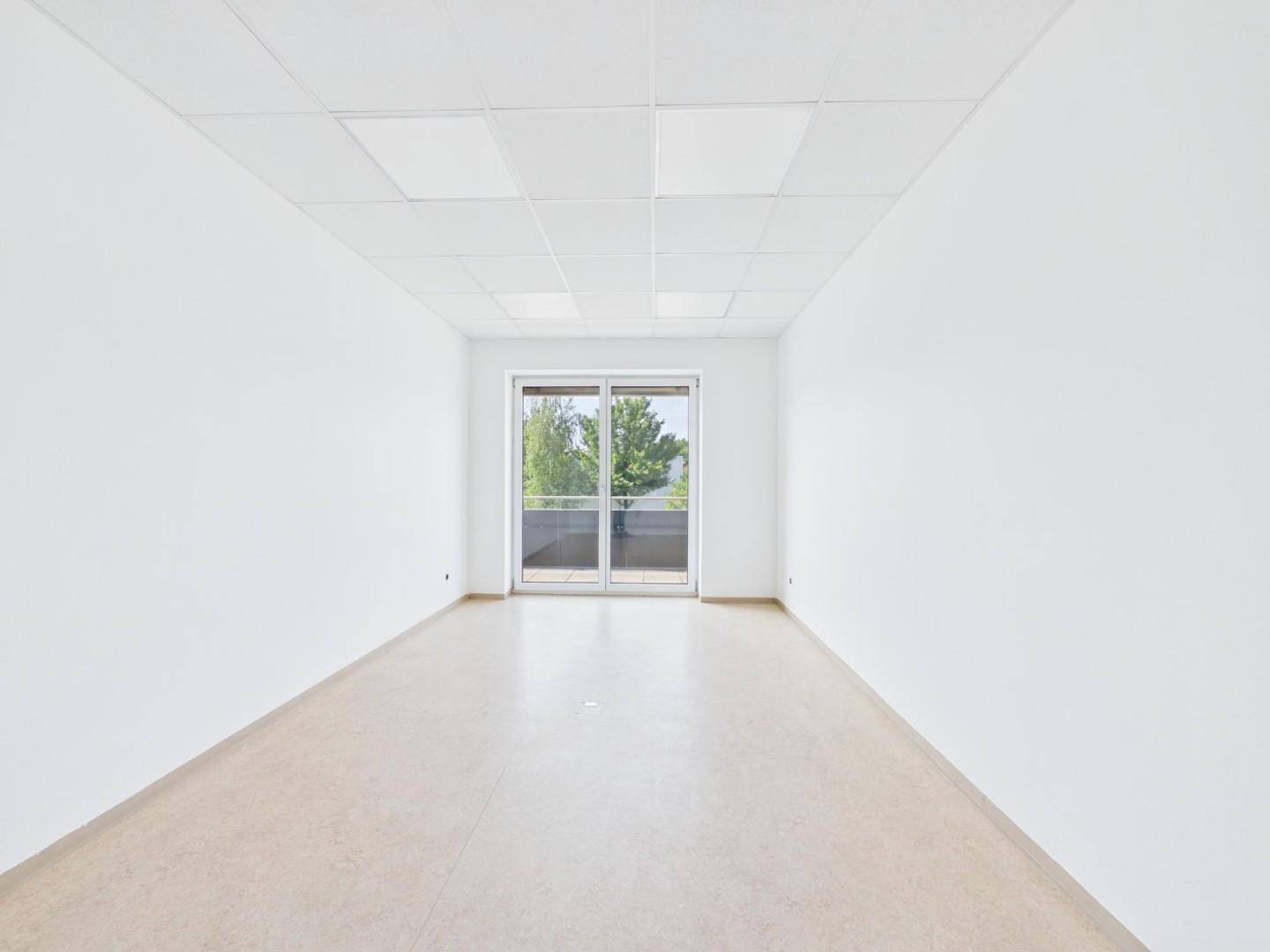 Langer, heller Raum mit einem großen Fenster am Ende, hellem Boden und Rasterdecke.