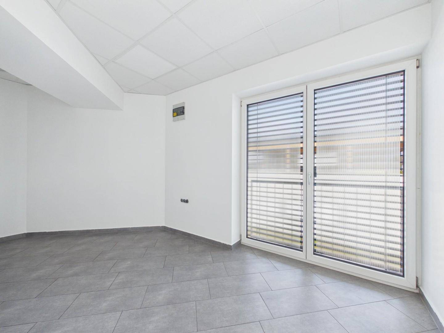 Leerer Raum mit grauen Fliesen, weißen Wänden und einem großen Fenster mit Jalousien.