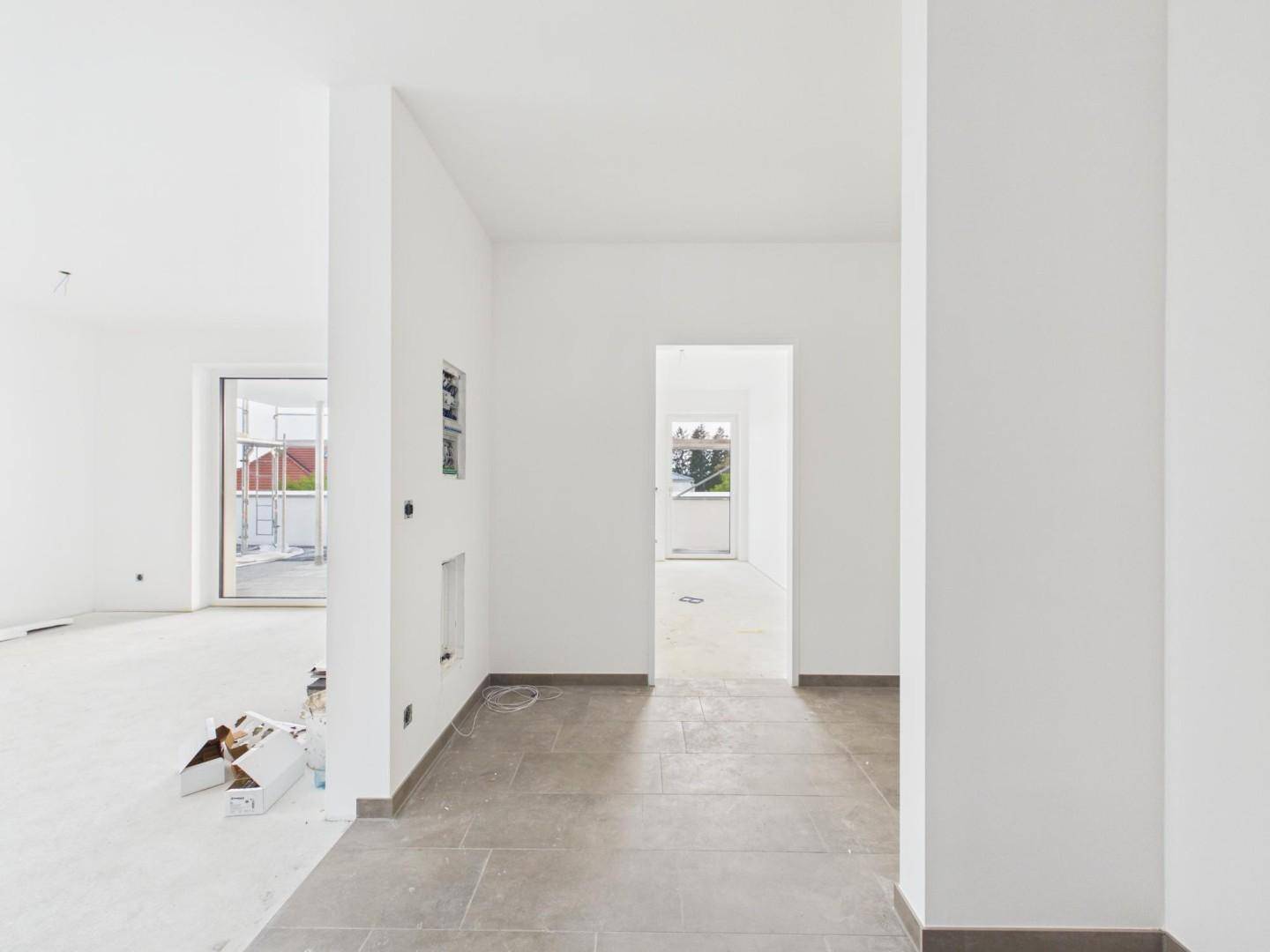 Heller Innenraum im Rohbau mit weißen Wänden und grauen Fliesenböden.